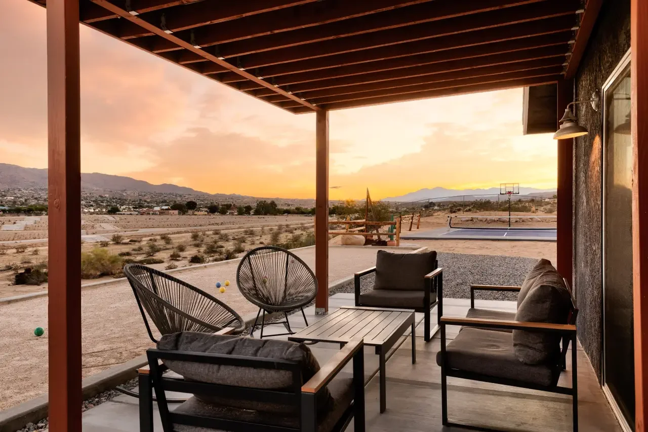 A covered patio with outdoor furniture overlooking a desert landscape at sunset, with a basketball hoop and a neighboring town visible in the distance.