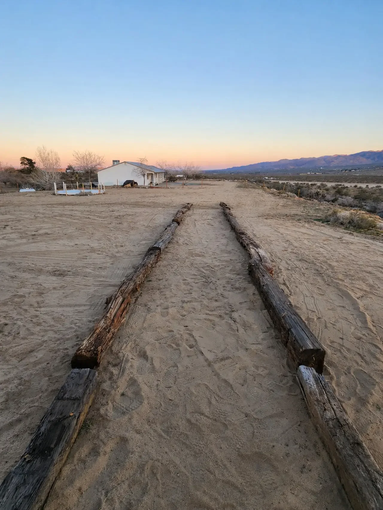 Wooden railroad ties laid out on sandy ground leading to a white house with trees and mountains in the background at sunset.