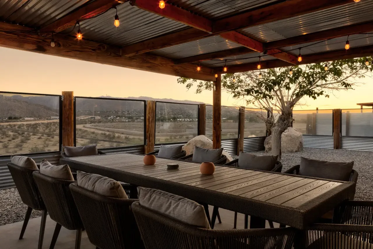 Outdoor dining area with a long wooden table and chairs, string lights hanging from a metal roof, a tree and rocks, and a desert landscape view at sunset.