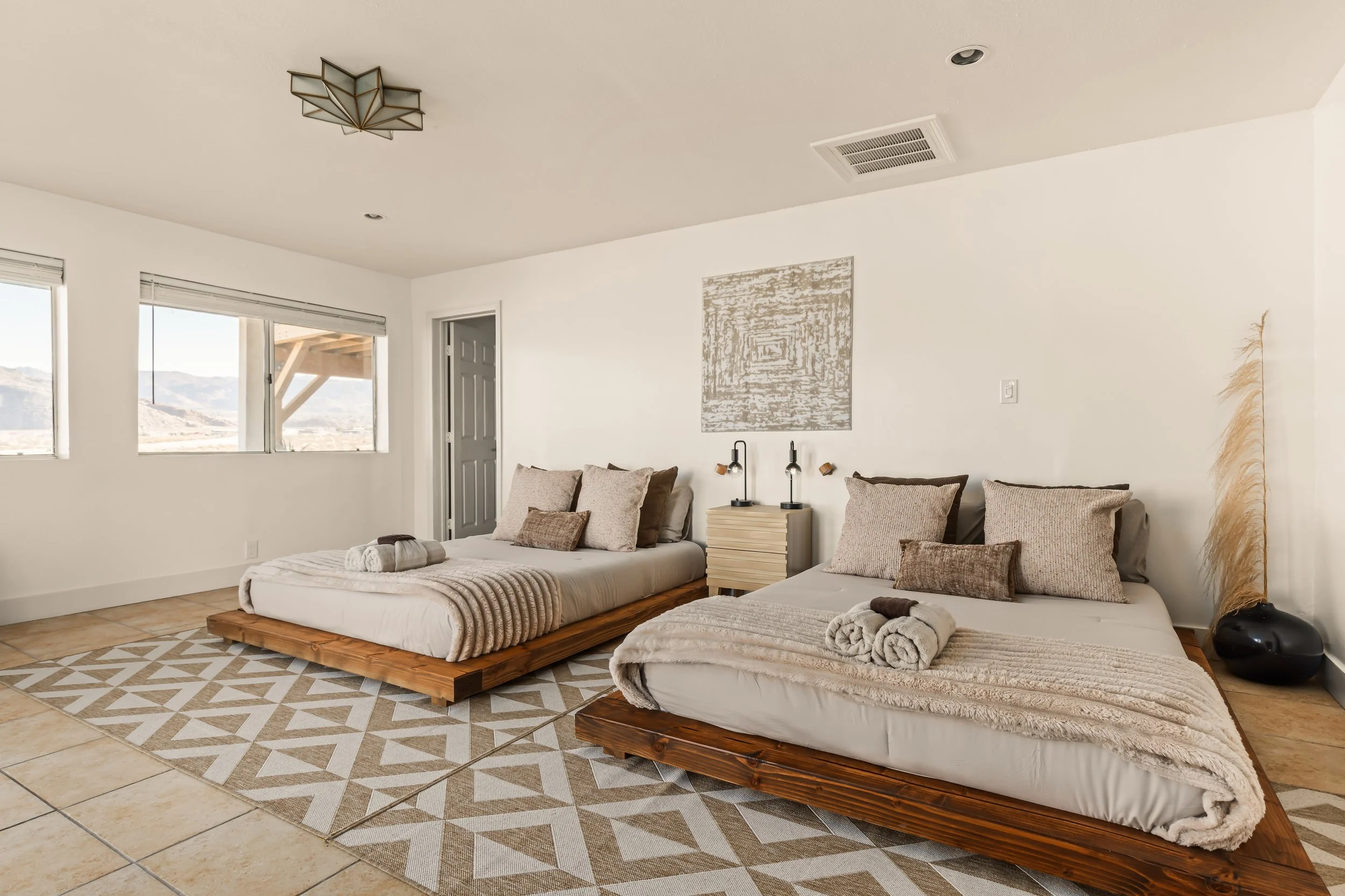 A bedroom with two beds on wooden frames, beige pillows, and rolled towels. The room has large windows with a view of mountains, a patterned rug, a nightstand with two black lamps, and a wall art piece. The ceiling has a modern light fixture and a ve