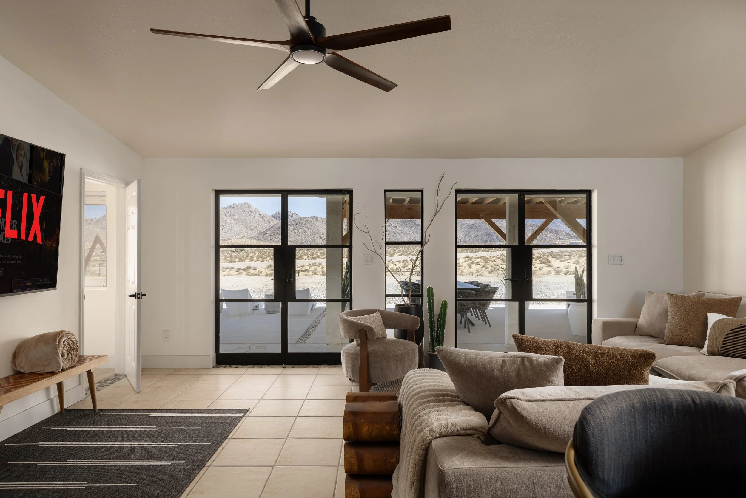 Living room with beige sofas, a beige chair, and a wooden coffee table in front of large black-framed sliding glass doors that reveal a desert landscape with mountains. White walls, a ceiling fan, and a television on the left wall.
