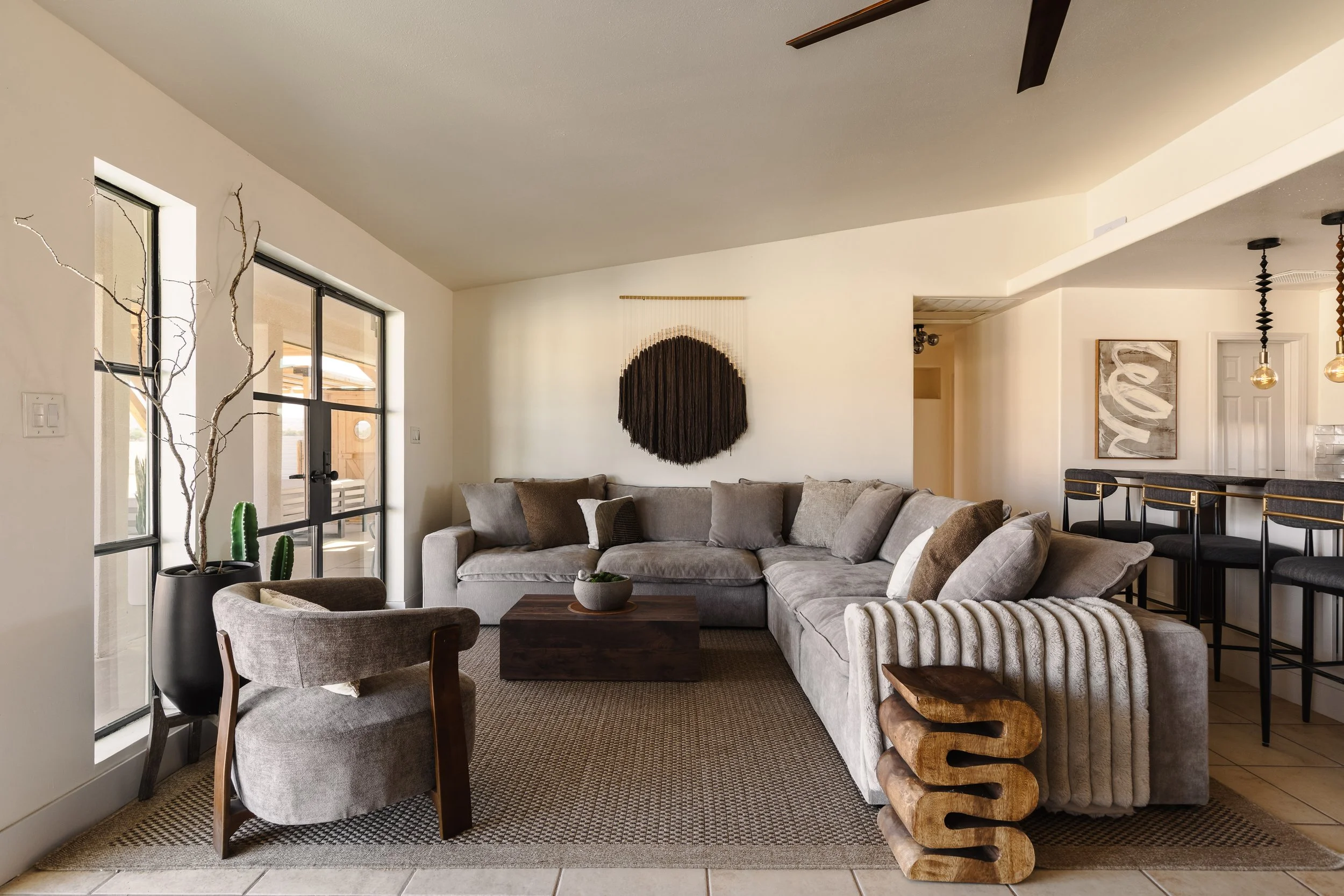 Living room with large gray sectional sofa, armchair, and wooden coffee table. Decor includes wall art, a hanging wall sculpture, and a potted plant near the window. Bright natural light from large windows.