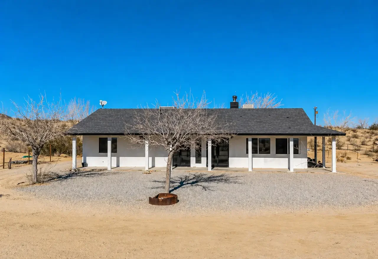 Single-story house in a desert landscape with leafless trees, gravel driveway, and a clear blue sky.