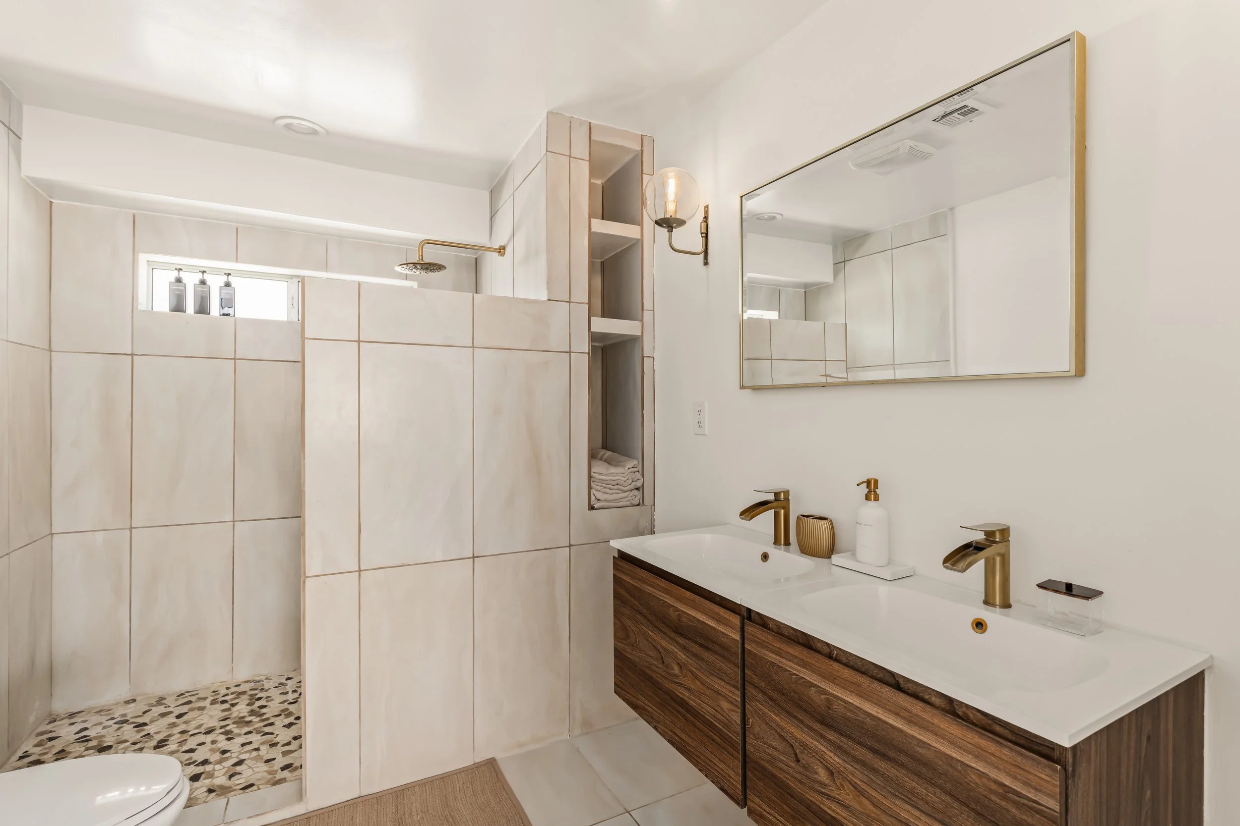 Modern bathroom with a shower area, a large mirror, double sinks with gold fixtures, wooden cabinets, and a small window in the shower.