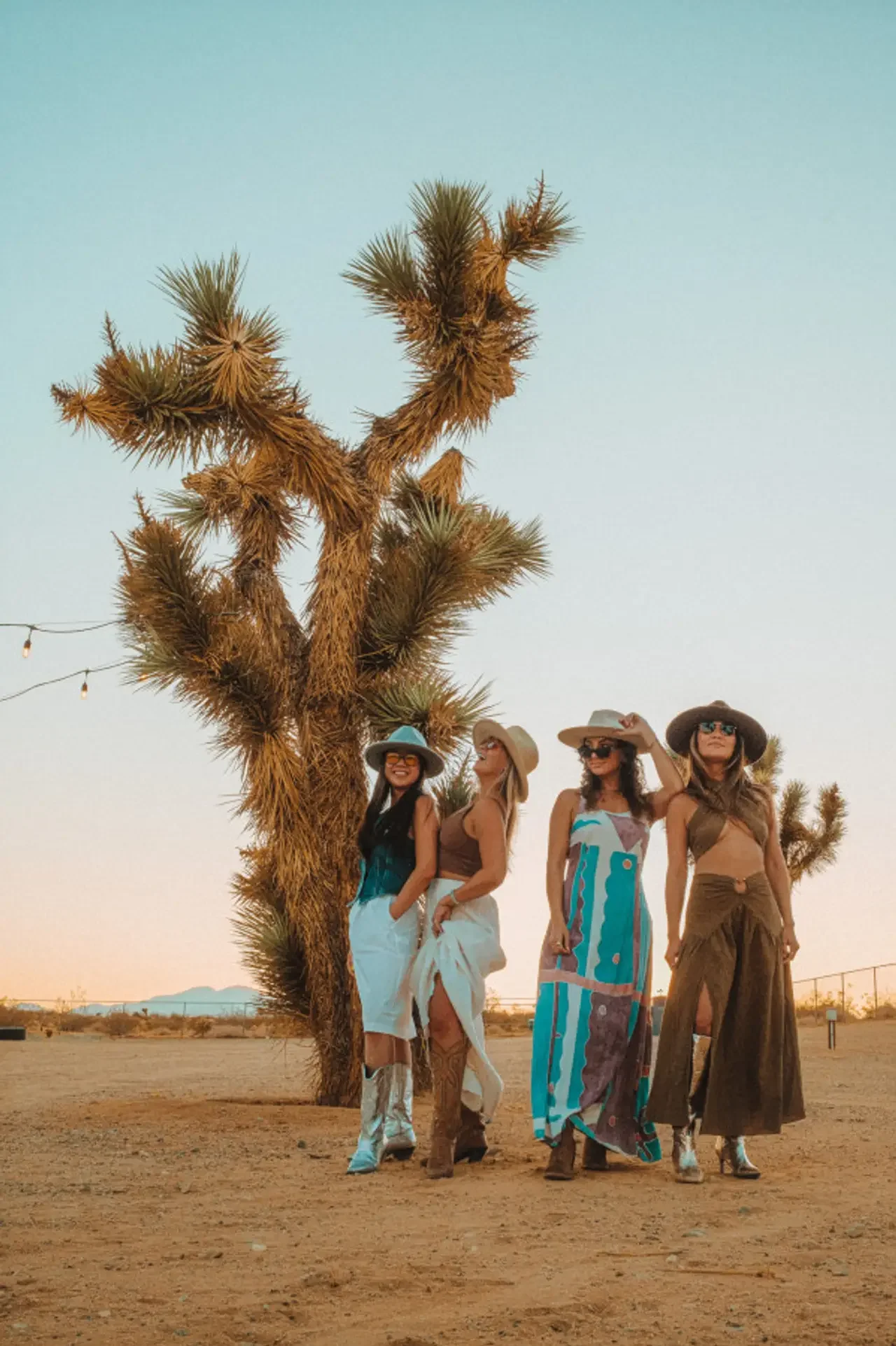 Four women standing beside a large desert tree, wearing straw hats and colorful summer dresses, in a desert landscape during sunset.