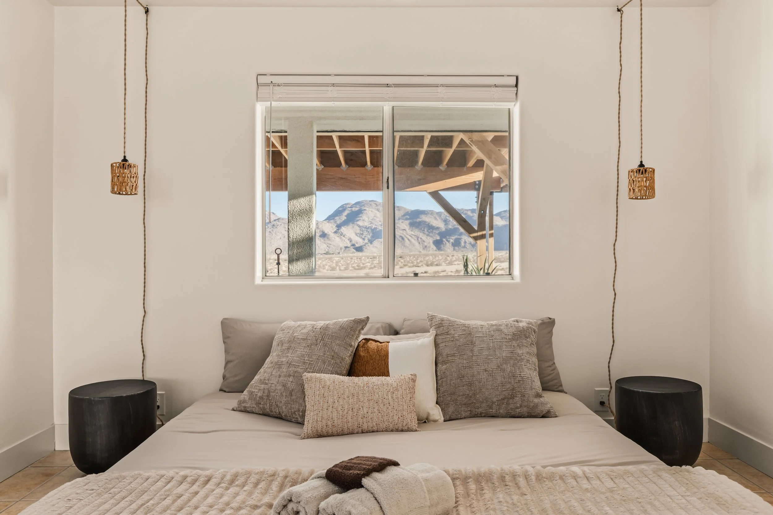 A bedroom with a large window showing mountains, decorated with hanging wicker lamps, a bed with multiple throw pillows, and a beige blanket.
