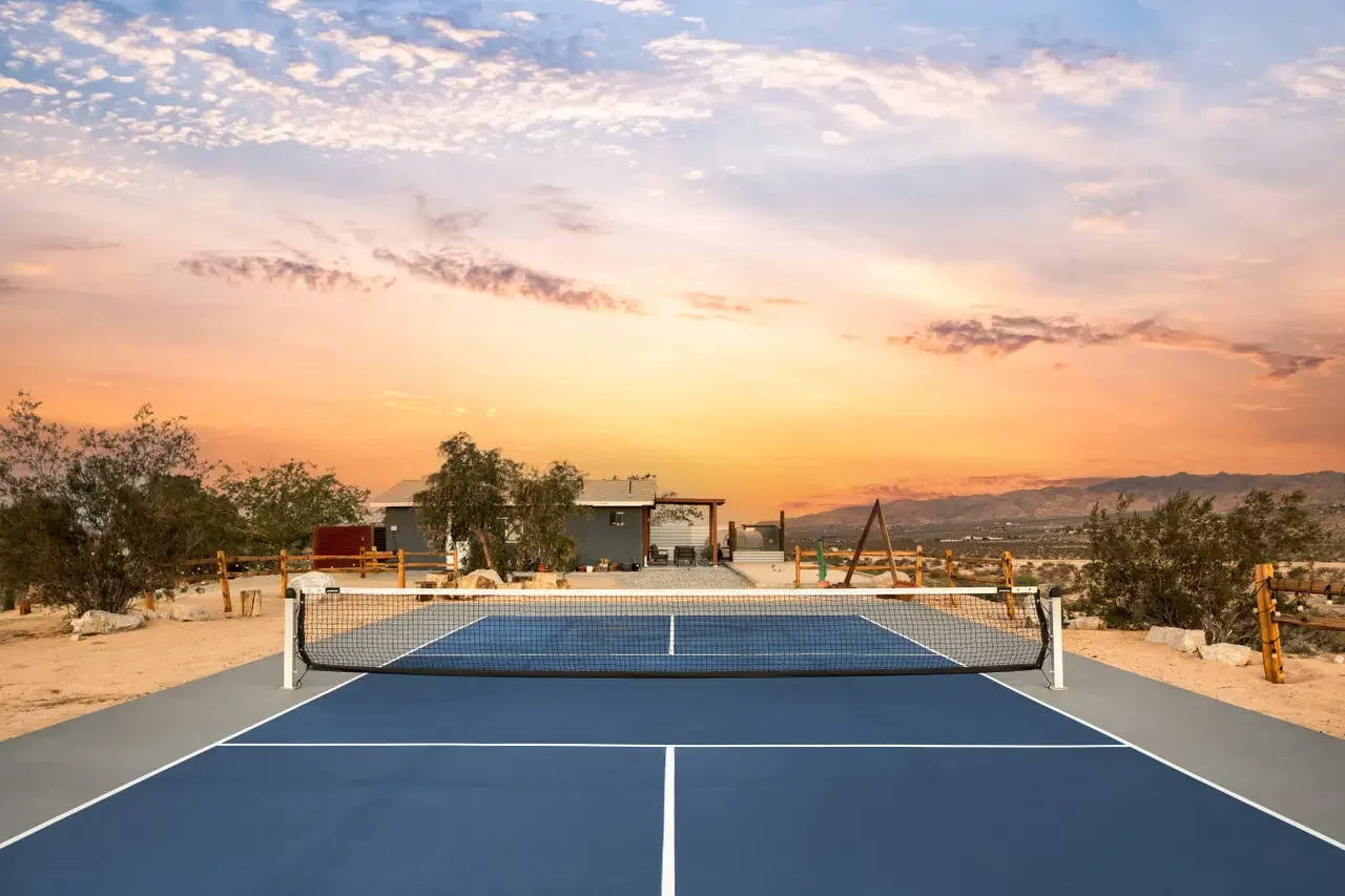 A blue tennis court in a desert-like area during sunset with a house, trees, and a play structure in the background.