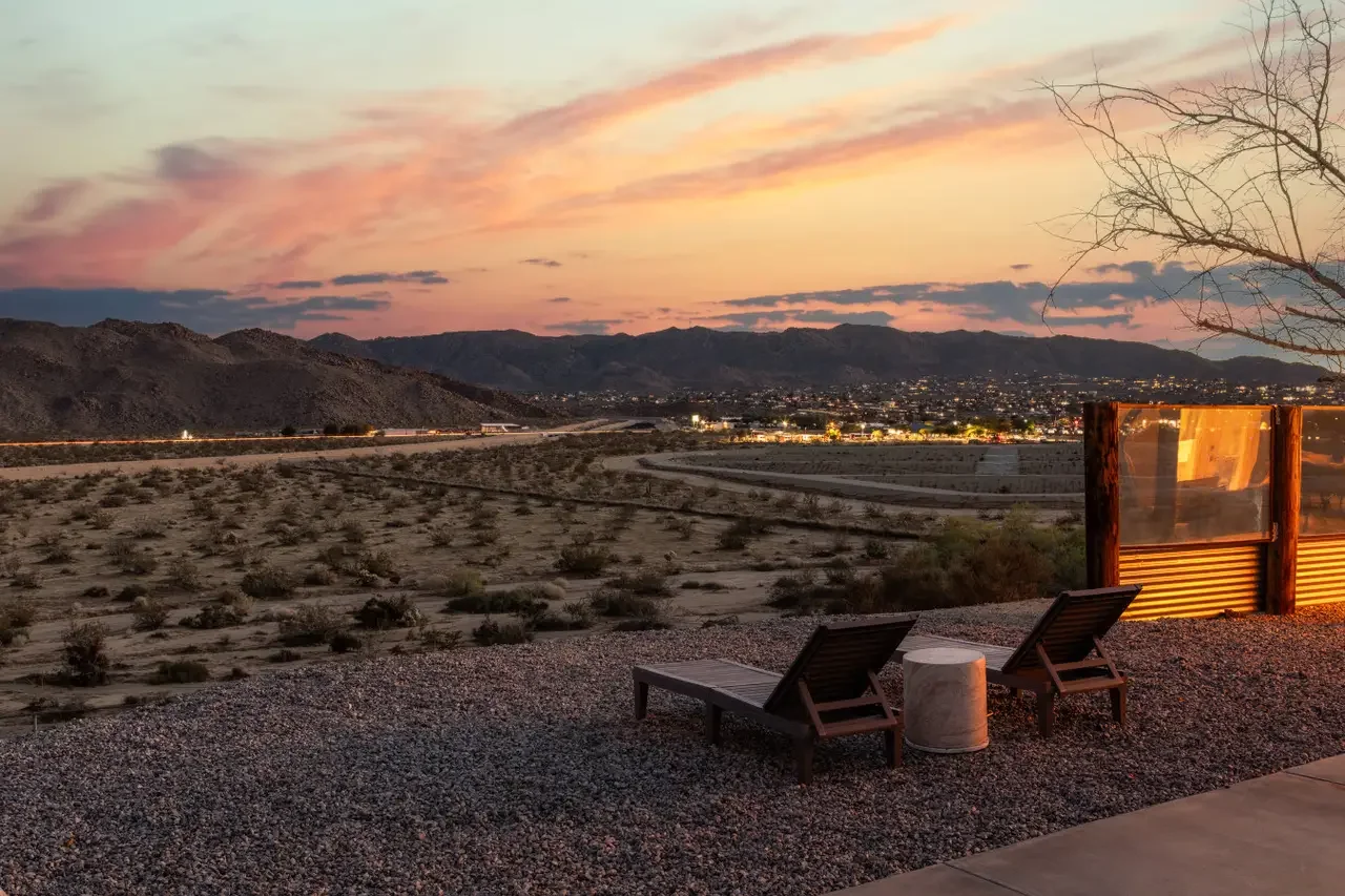 Two lounge chairs and a small round table on gravel patio overlooking a desert landscape with mountains at sunset, city lights in the distance, and a partly cloudy sky.
