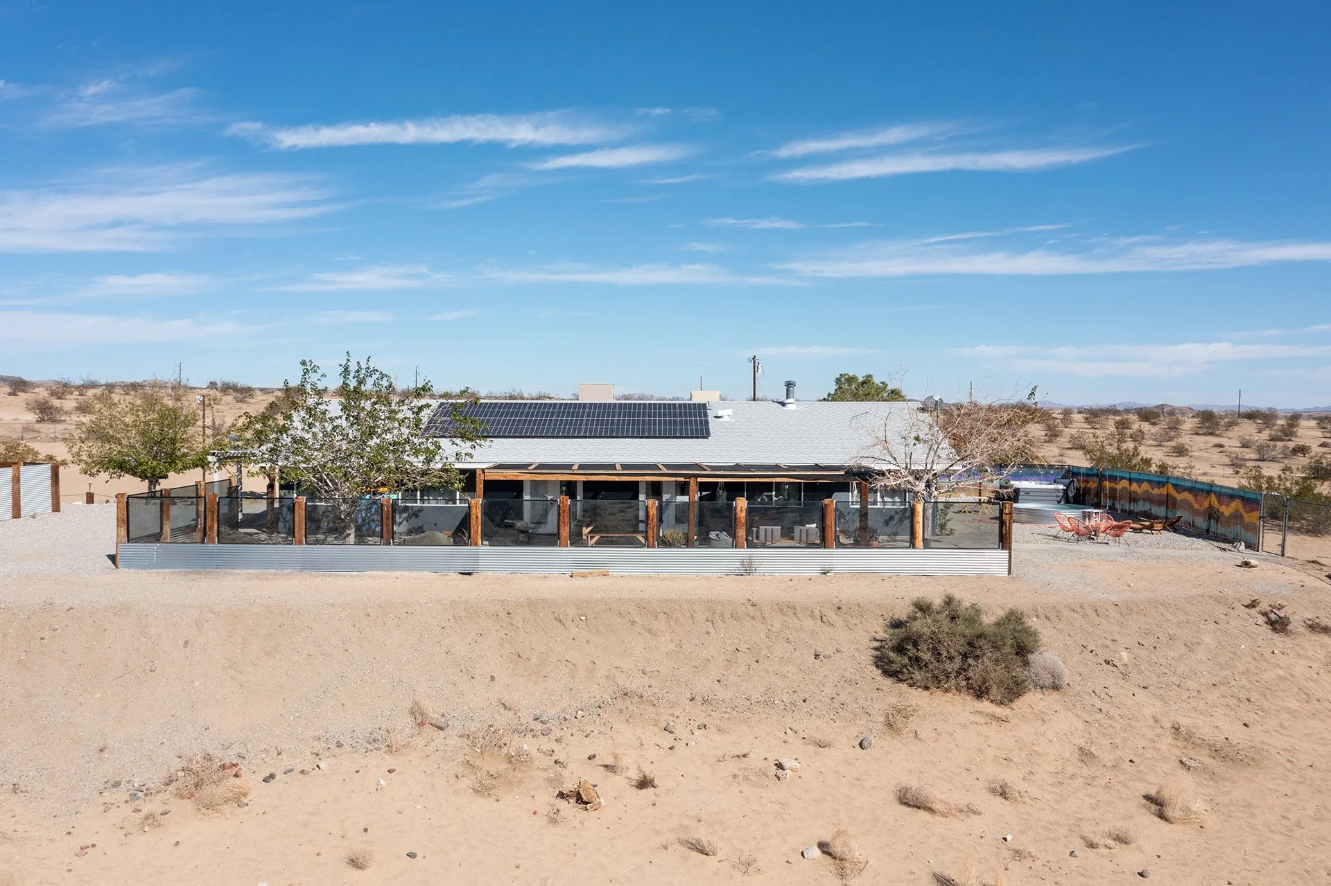 A house in a desert landscape with solar panels on the roof, surrounded by a fenced yard with trees and outdoor seating, under a blue sky with clouds.
