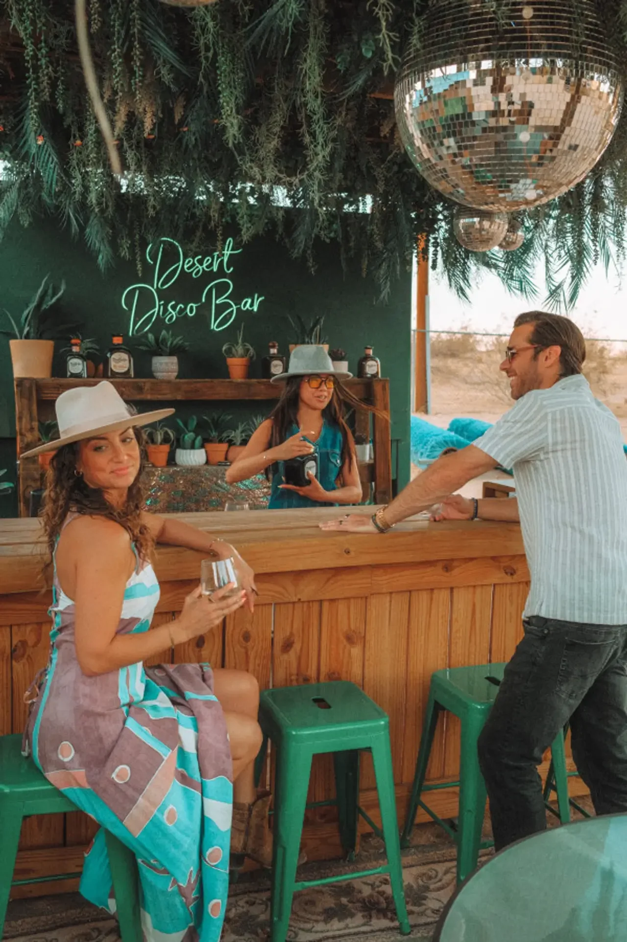 Three people at a bar with a green wall that reads 'Desert Disco Bar' and potted plants, one woman sitting on a green stool holding a glass, another woman behind the bar wearing a hat and glasses, and a man standing and smiling.