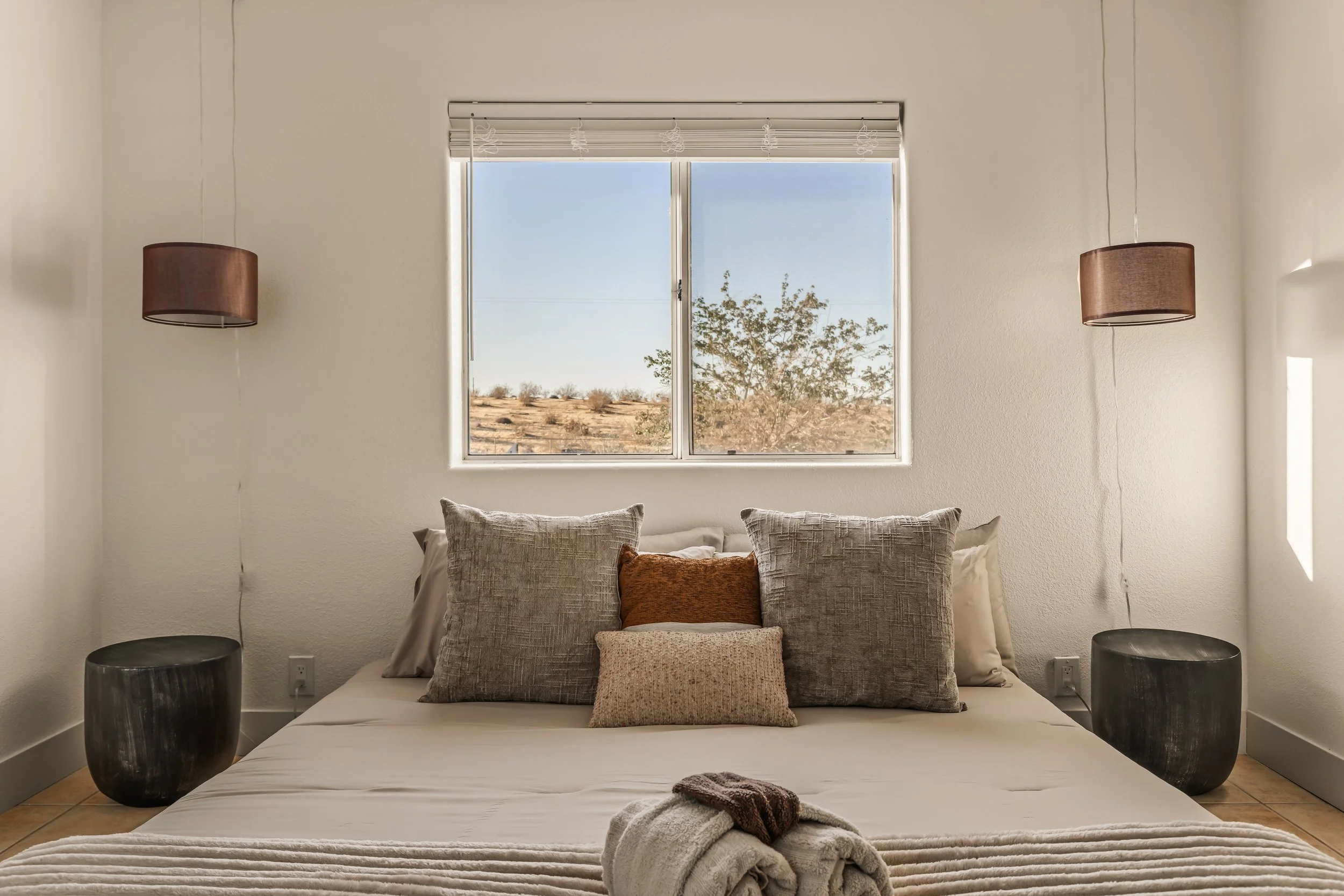 A bedroom with a bed that has multiple pillows and a blanket, two black side tables, a window with blinds showing an outdoor landscape, and brown pendant lights hanging on each side.