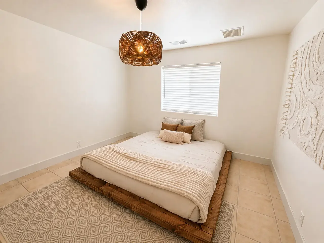 A minimalist bedroom with a low wooden bed frame, beige bedding, and decorative pillows, a window with blinds, a textured wall art on the right, a woven pendant light hanging from the ceiling, and tiled flooring with a patterned rug.