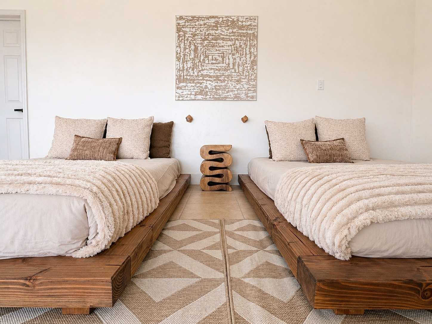A minimalistic bedroom with two beds on wooden platforms, beige and brown pillows, plush beige blankets, a geometric-patterned rug, and an abstract wall art piece.