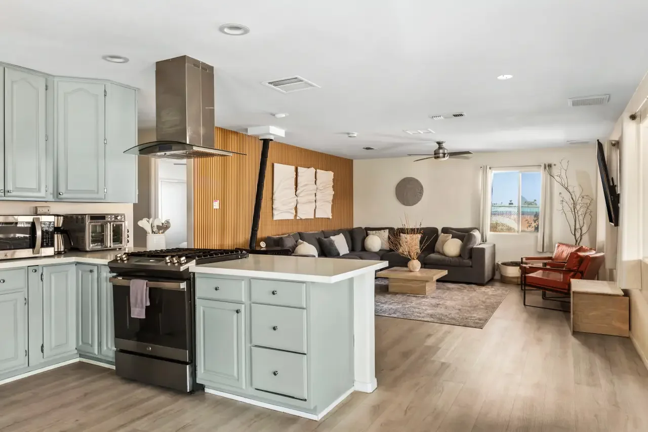 Open-concept kitchen and living room with light blue cabinets, stainless steel appliances, black sectional sofa, red armchair, wall-mounted TV, wooden coffee table, and large window with curtains.