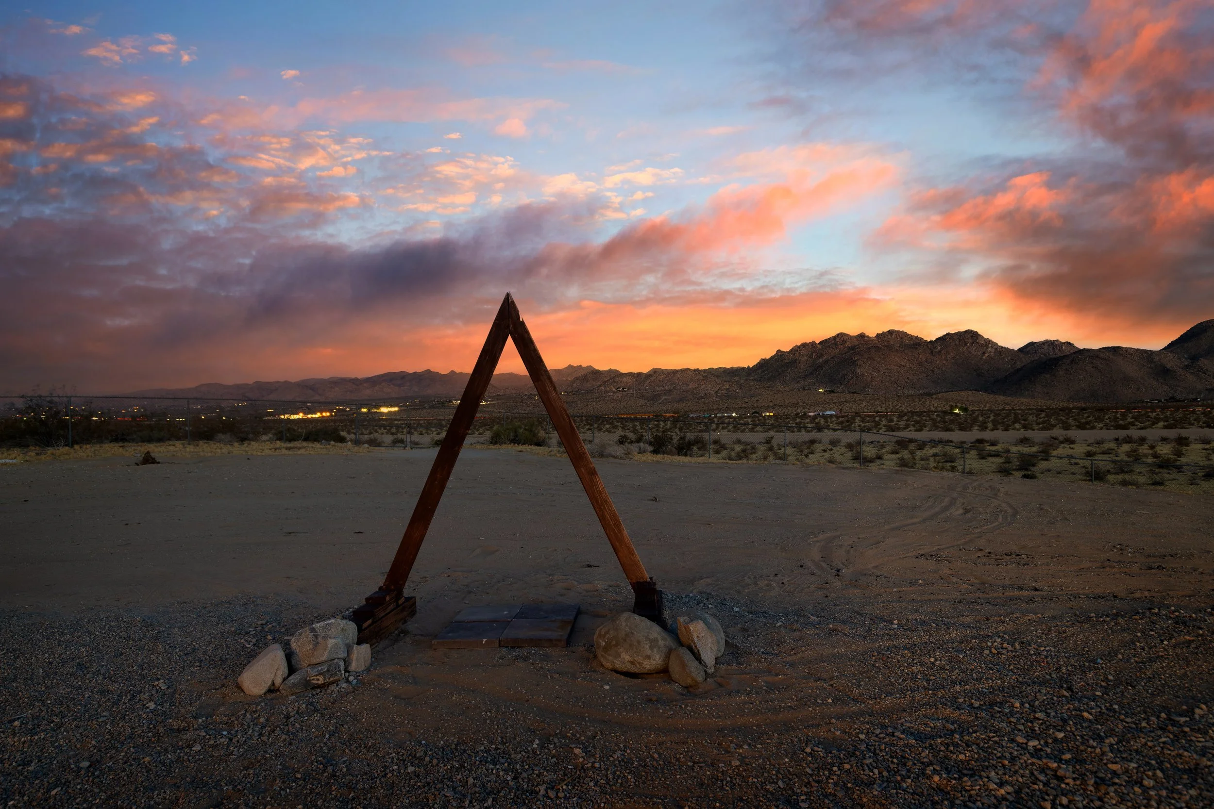 A desert landscape at sunset with mountains in the distance, colorful clouds in the sky, and a wooden A-frame structure in the foreground surrounded by rocks.