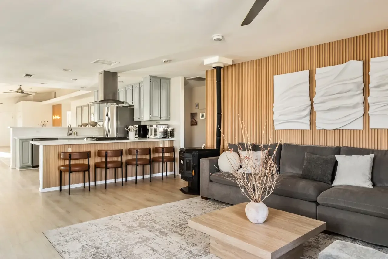 Open-concept living room and kitchen with a gray sectional sofa, wooden coffee table with a white vase and dried branches, wood-paneled accent wall with three white textured art pieces, and kitchen with gray cabinets, a island with bar stools, stainl