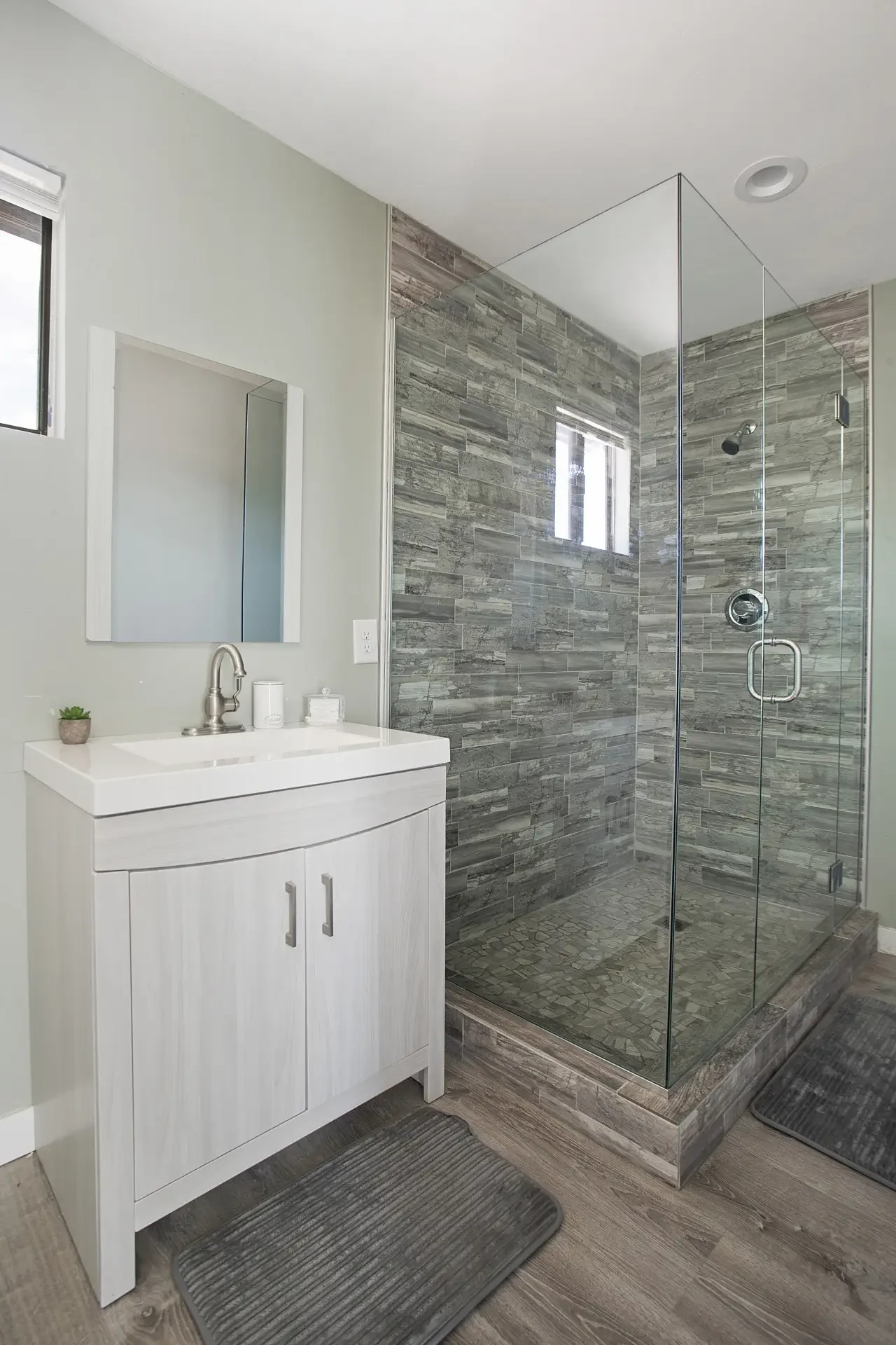 Modern bathroom with a white vanity, rectangular mirror, small potted plant, soap dispenser, glass shower enclosure with gray stone tiles, small window, brown wooden floor, and dark gray bath mats.
