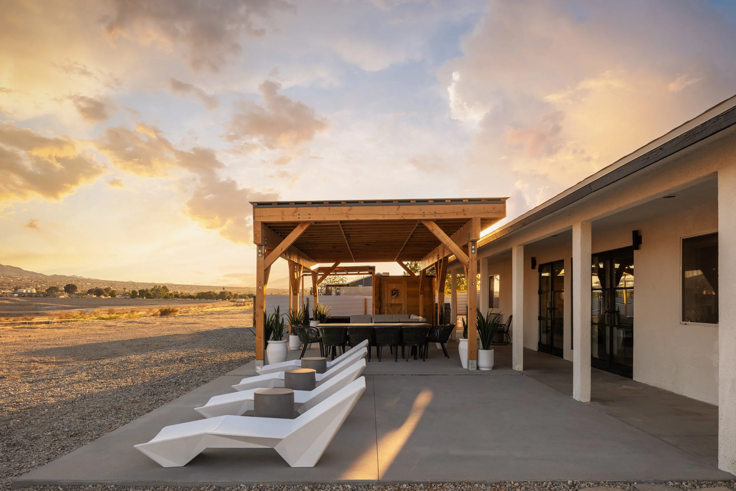 A backyard patio with white lounge chairs, a dining table with chairs underneath a wooden pergola, potted plants, and a view of a desert landscape at sunset.