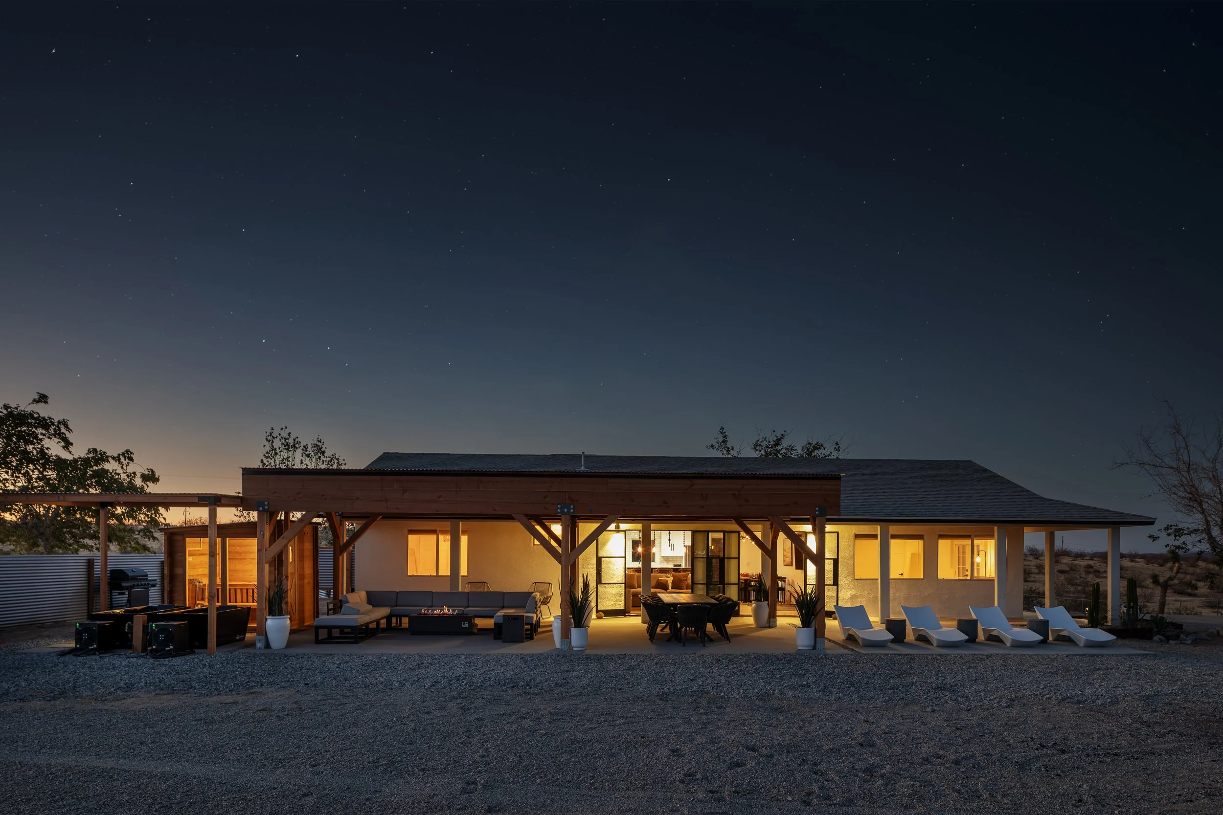 A modern house illuminated with warm interior lighting under a starry night sky, with an outdoor patio area featuring sofas, chairs, lounge chairs, and desert plants, and a gravel yard in the foreground.