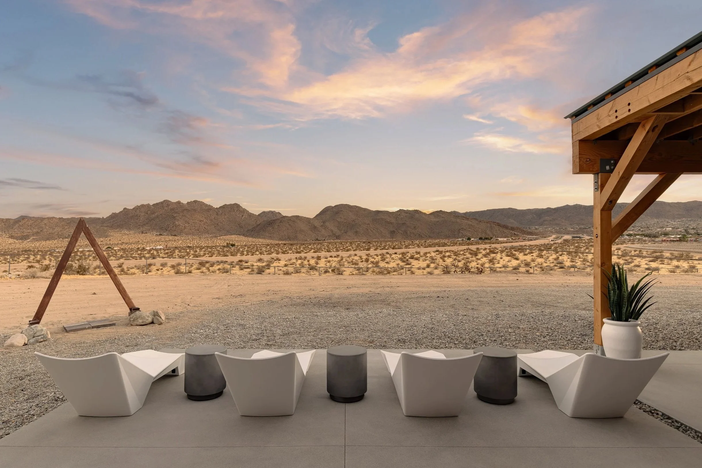 A desert landscape with a mountain range in the background during sunset, with a modern outdoor seating area and a partially visible wooden structure on the right.