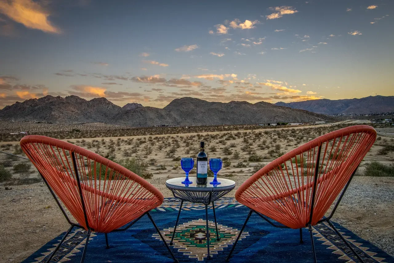 A desert landscape during sunset with mountains and a partly cloudy sky, featuring two orange wire chairs facing a small round table with a bottle of wine and two blue wine glasses on a colorful rug.