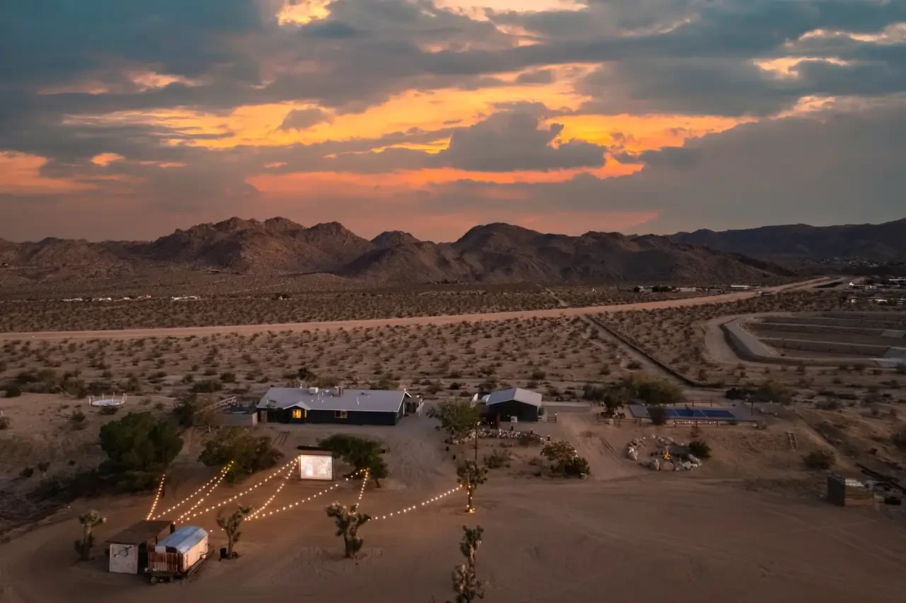 Desert landscape at sunset with mountains in the background, a few scattered buildings, and string lights in the foreground.