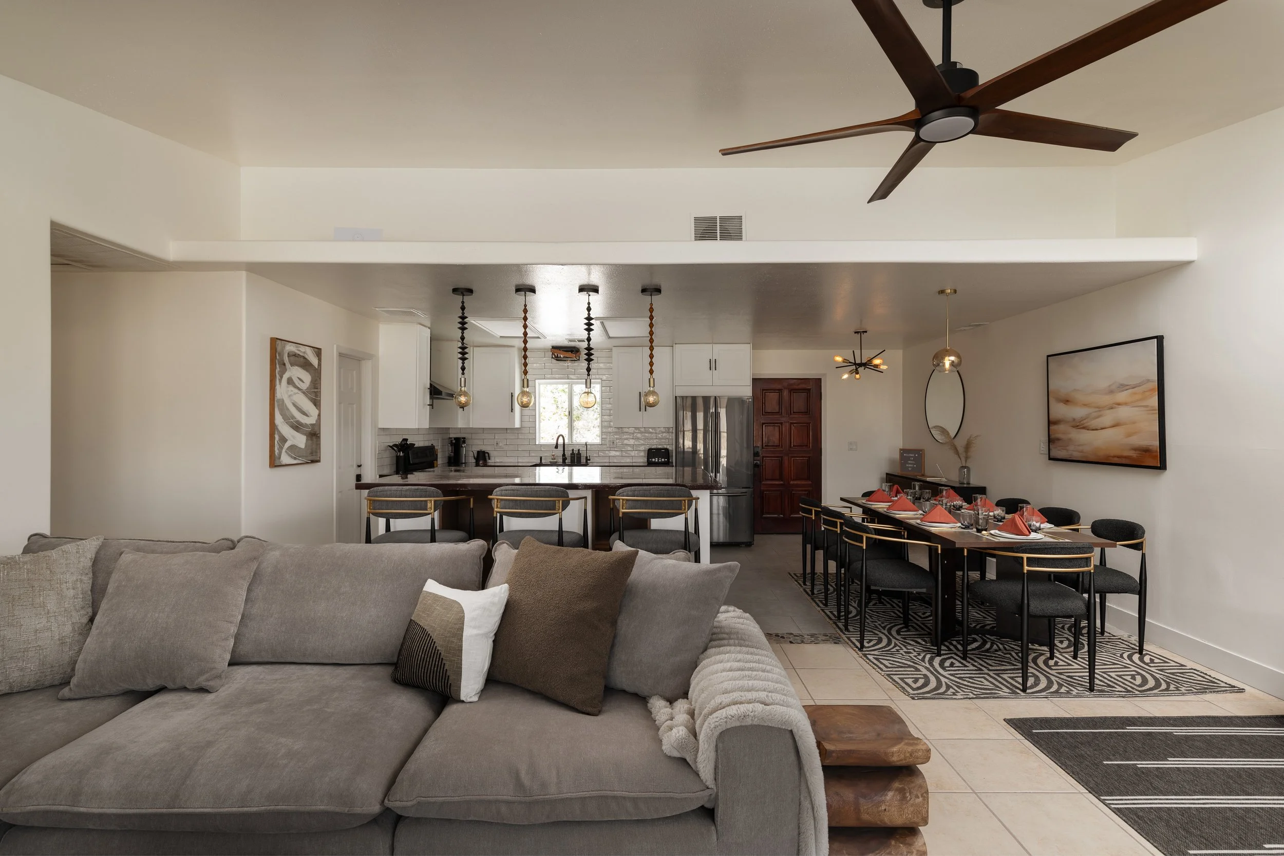 Open-concept living and dining area with a kitchen in the background. The living room has a gray sofa with pillows and a blanket. The dining area has a long black table with red napkins, glasses, and black chairs. The kitchen features white cabinets,