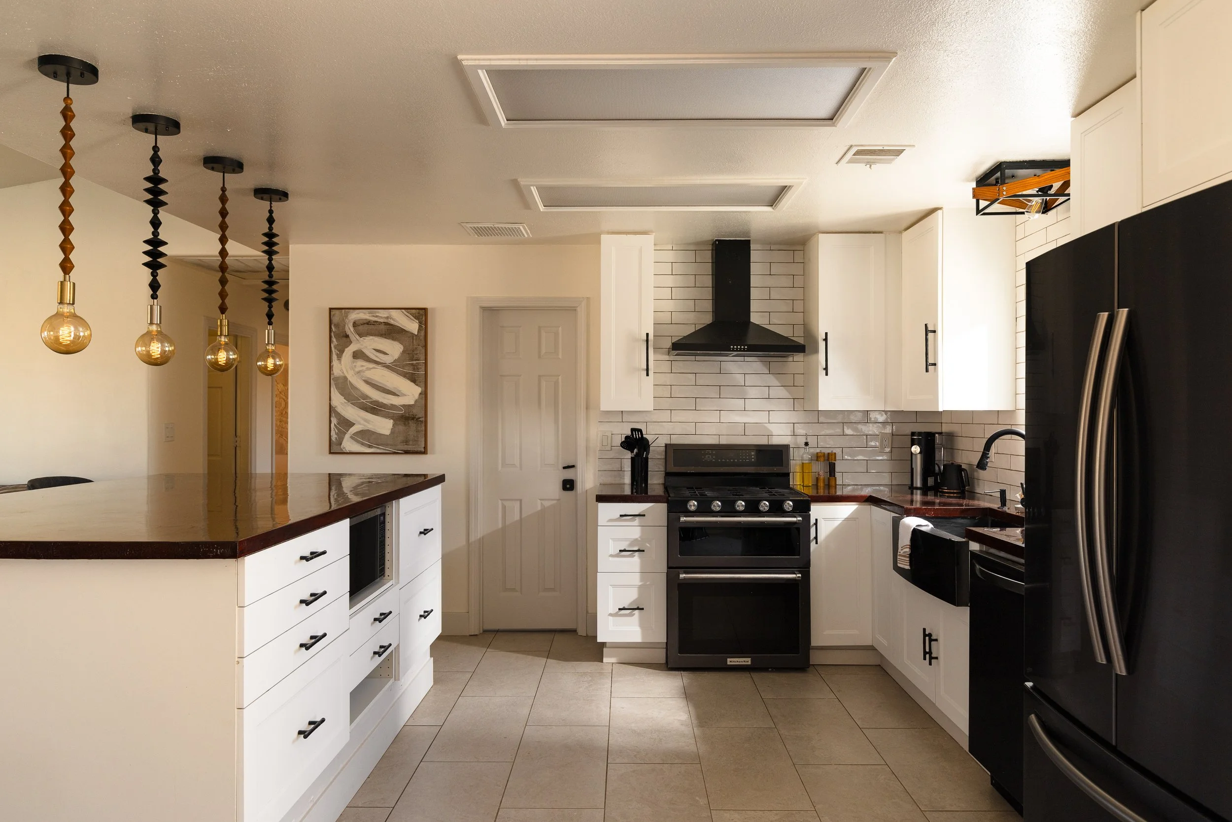 A modern kitchen with white cabinets, black appliances, a stainless steel refrigerator, and a white tiled backsplash. Pendant lights hang above a white island with a dark wooden countertop. There is artwork on the wall and various kitchen utensils an
