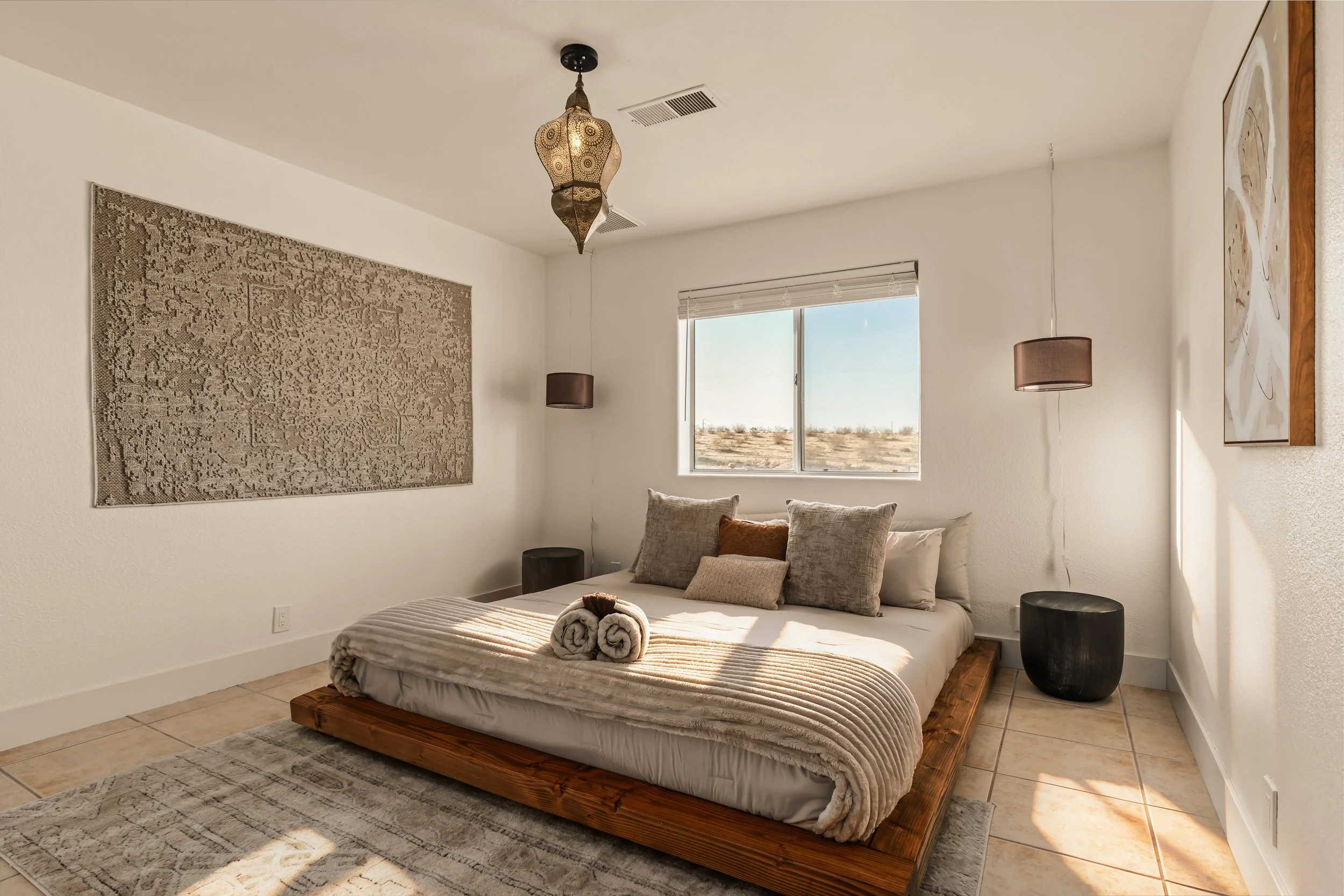A minimalist bedroom with a low wooden platform bed decorated with neutral pillows and rolled towels. The room features a large window letting in natural light, with desert landscape visible outside. The walls are adorned with textured art and a larg