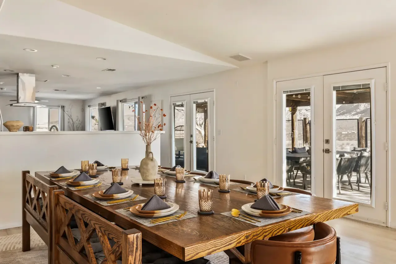 Dining room with a long wooden table set with black and gold plates and napkins, glassware, and a flower vase; large glass doors and windows in the background.