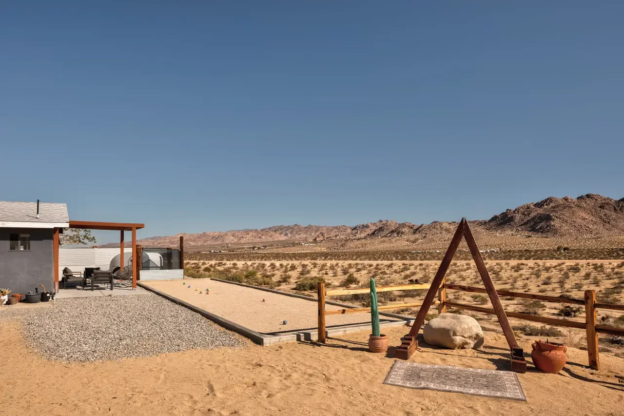 Desert landscape with a fenced backyard, a small building, outdoor seating area, and a gravel playing area for outdoor games.
