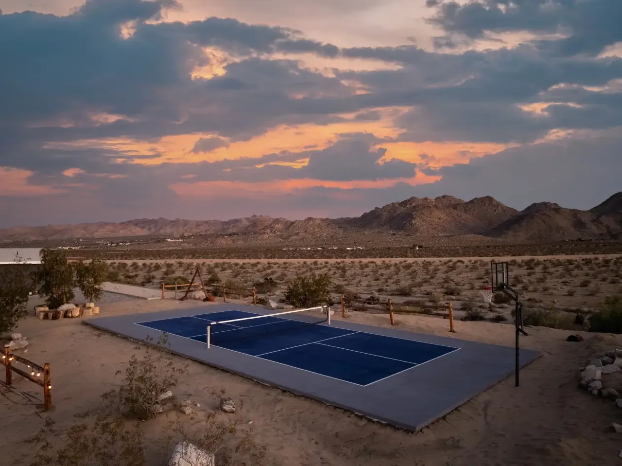 A desert landscape at sunset with mountains in the background, featuring a mounted basketball hoop and a portable tennis court in the foreground.