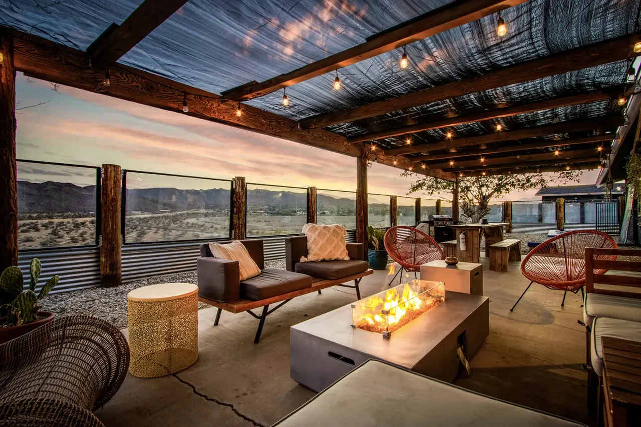 Outdoor patio with string lights, wooden beams, and a fire table, offering a desert mountain view at sunset.