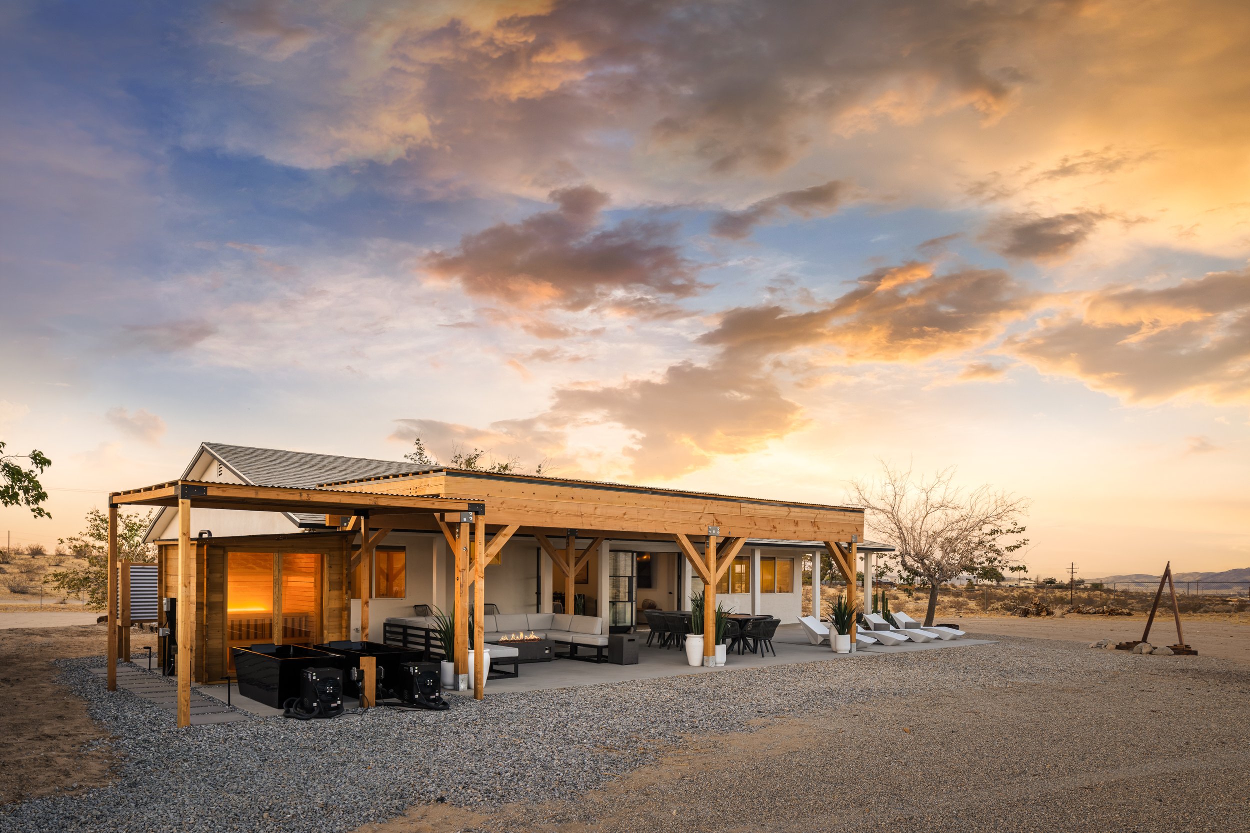 Modern house with outdoor patio under a colorful sunset sky, surrounded by desert landscape and sparse trees.