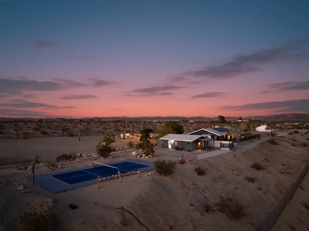 A sunset view of a desert landscape with a house, a fenced yard, and a basketball court. The sky has shades of pink, purple, and blue with scattered clouds.
