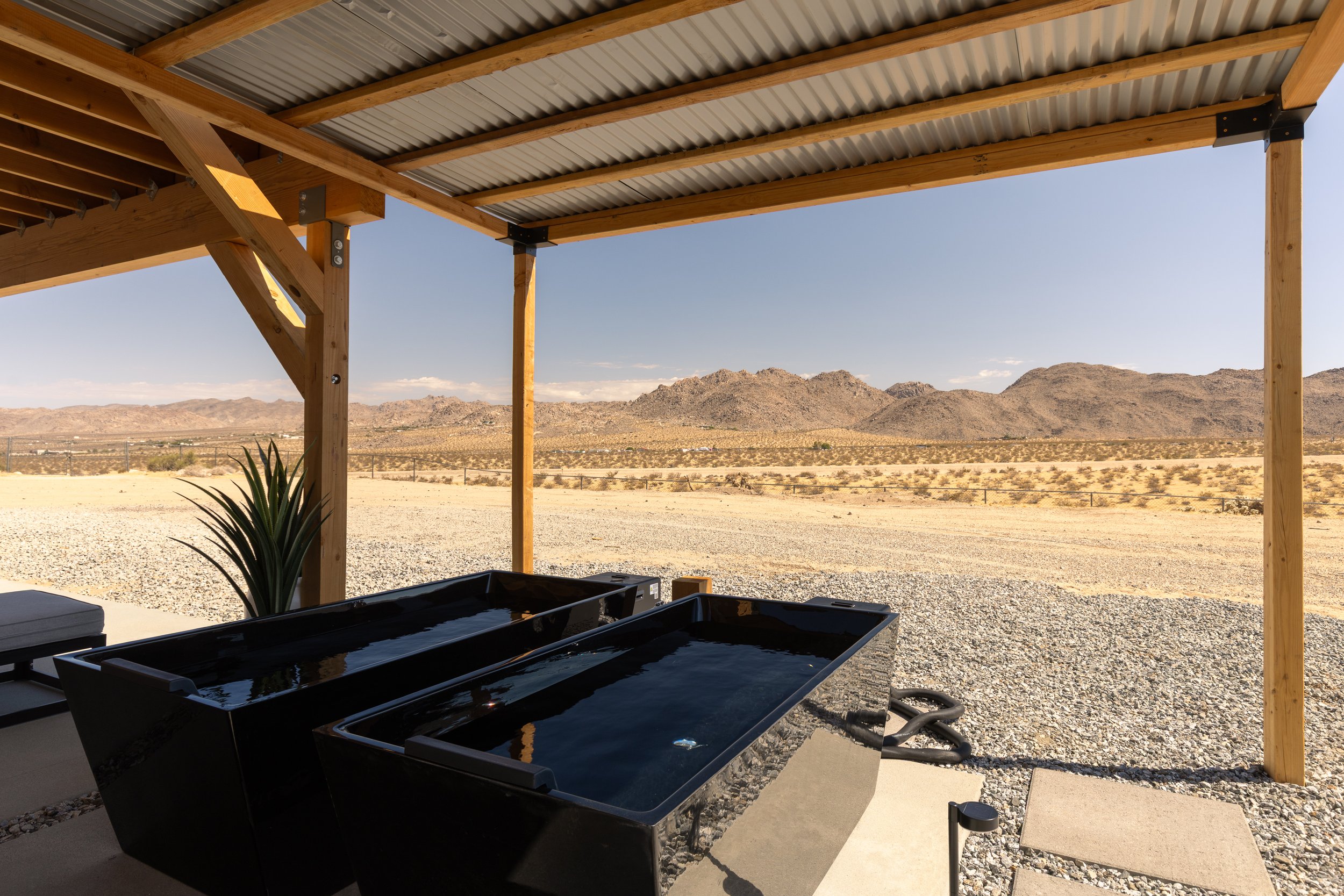 Outdoor shade structure with wooden beams and corrugated metal roof, overlooking a desert landscape with mountains, under a clear blue sky.