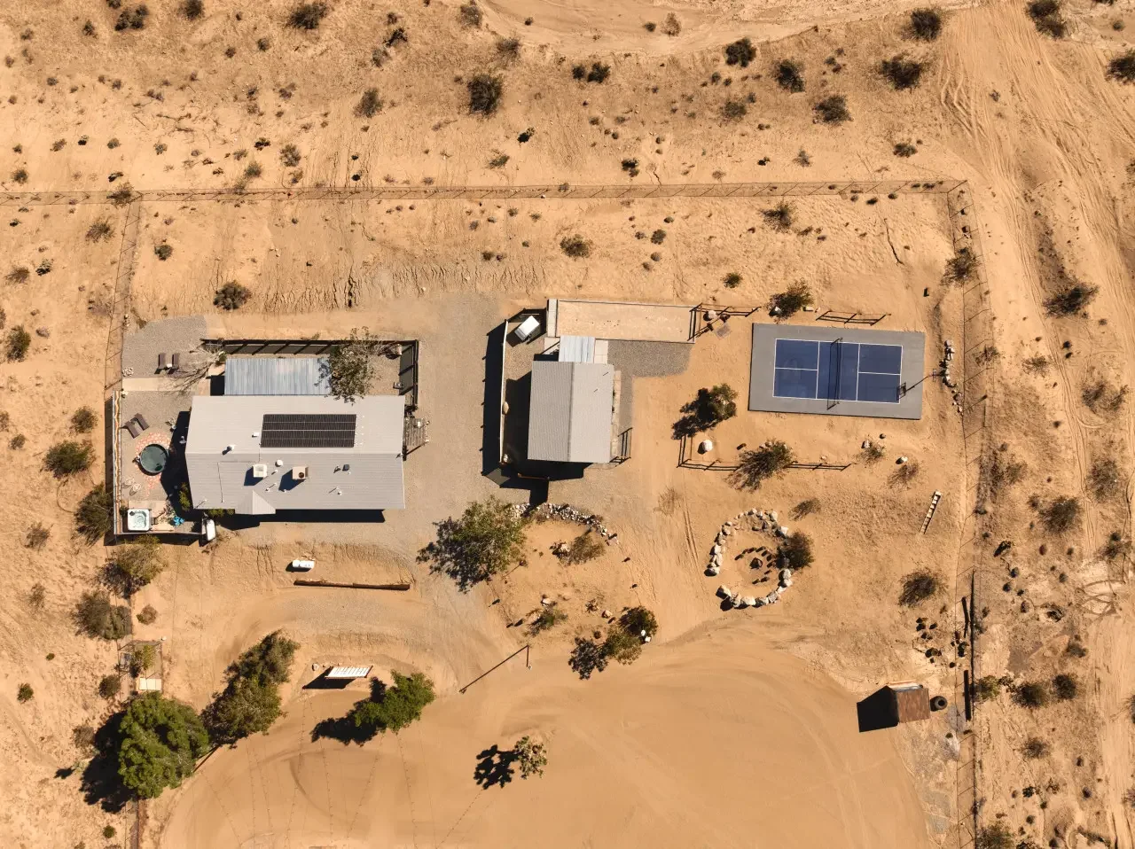 Aerial view of a desert property featuring a house with solar panels, a hot tub, a shed, a fenced tennis court, trees, a circular rock formation, and dirt paths surrounded by sparse desert vegetation.