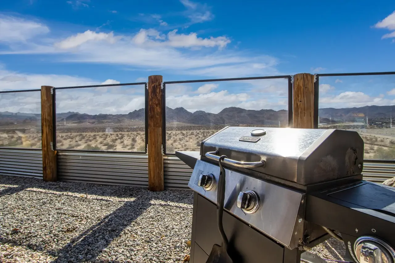 A stainless steel barbecue grill on a gravel surface with a scenic desert landscape and mountains in the background, under a partly cloudy sky.