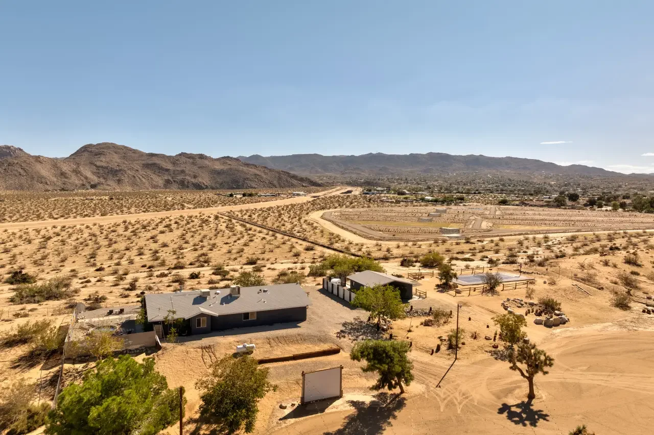 Aerial view of a desert landscape with a house, some trees, and distant mountains under a clear blue sky.