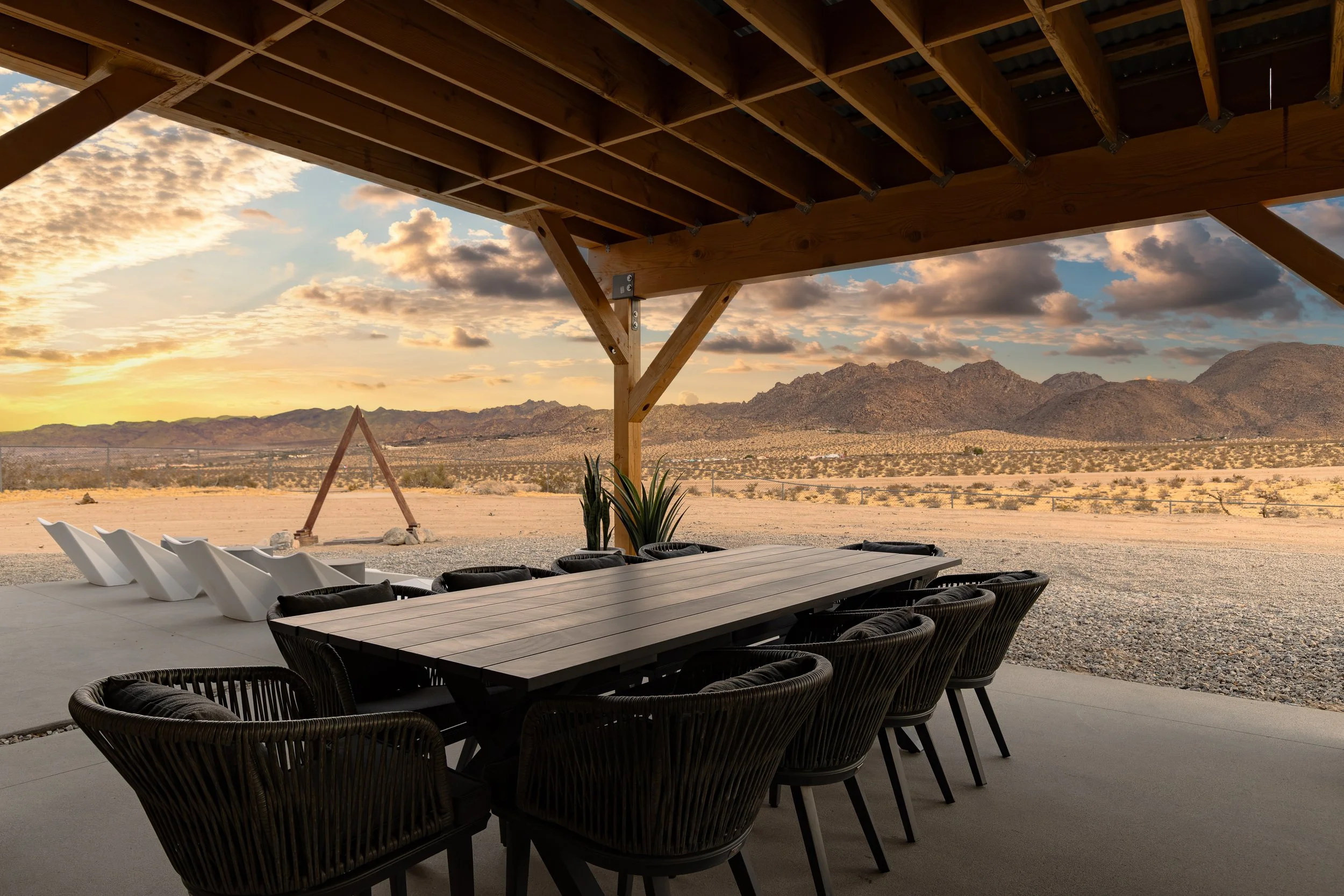 A modern outdoor patio with a long rectangular table and chairs, overlooking a desert landscape with mountains in the distance during sunset, with the patio partially covered by a wooden structure.