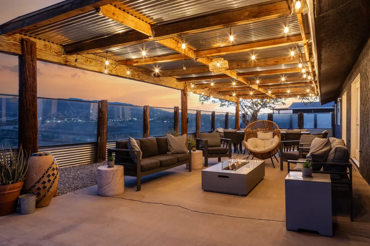 Outdoor patio with string lights, seating area with couches, chairs, and a fire pit, overlooking a cityscape at sunset.