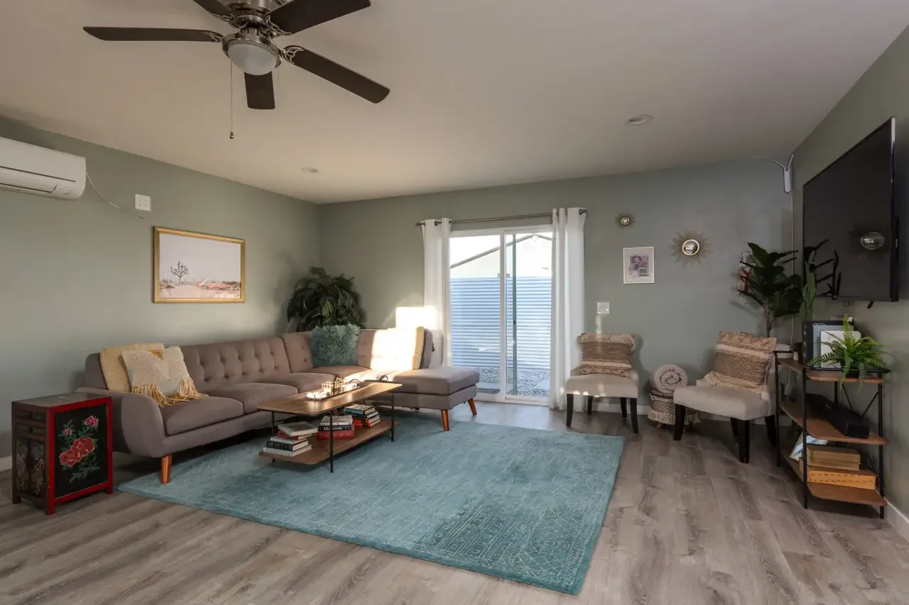 Living room with gray sectional sofa, two beige chairs, a teal area rug, potted plants, wall art, and a mounted flat-screen TV.