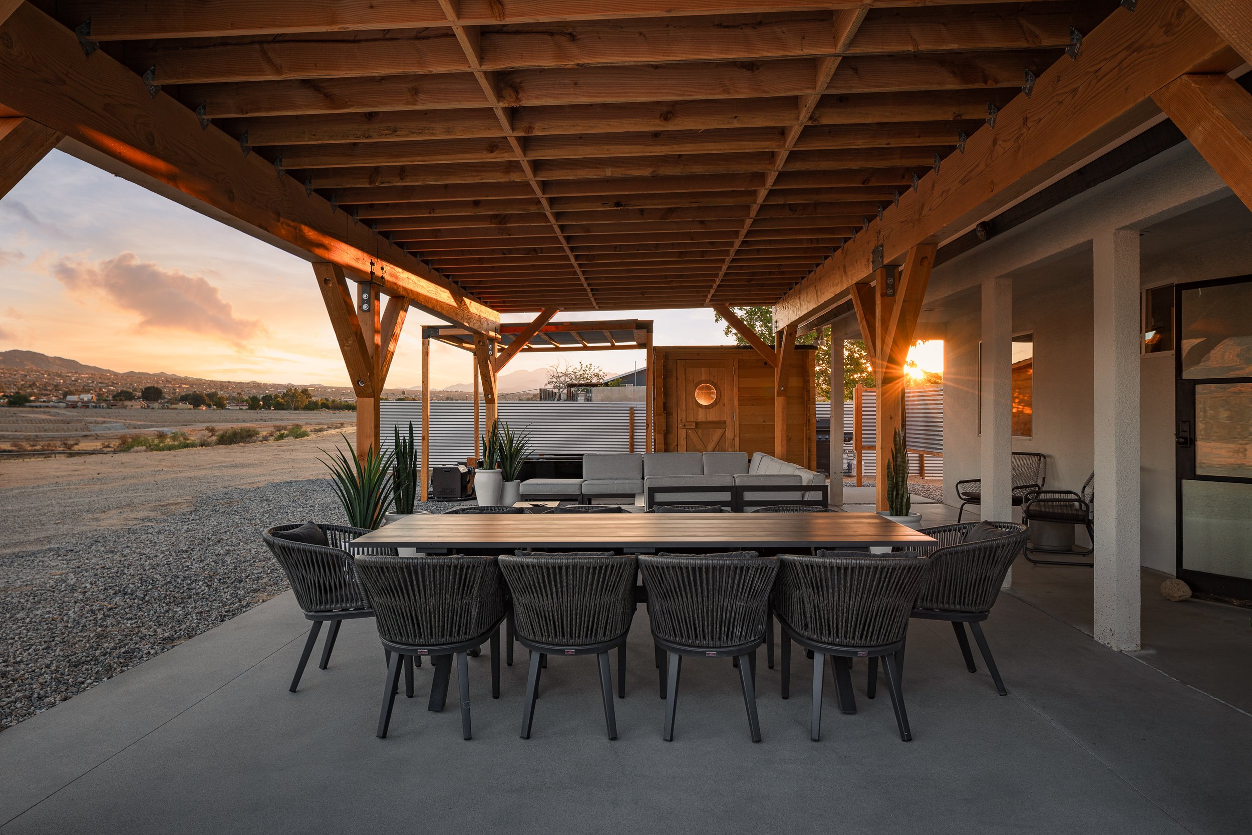 Outdoor patio with a wooden pergola, modern dining table with black chairs, a white sectional sofa, and potted plants at sunset, overlooking a landscape.