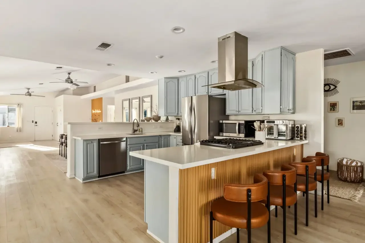 Open-concept kitchen with light blue cabinets, white countertops, stainless steel appliances, and a breakfast bar with brown leather chairs.