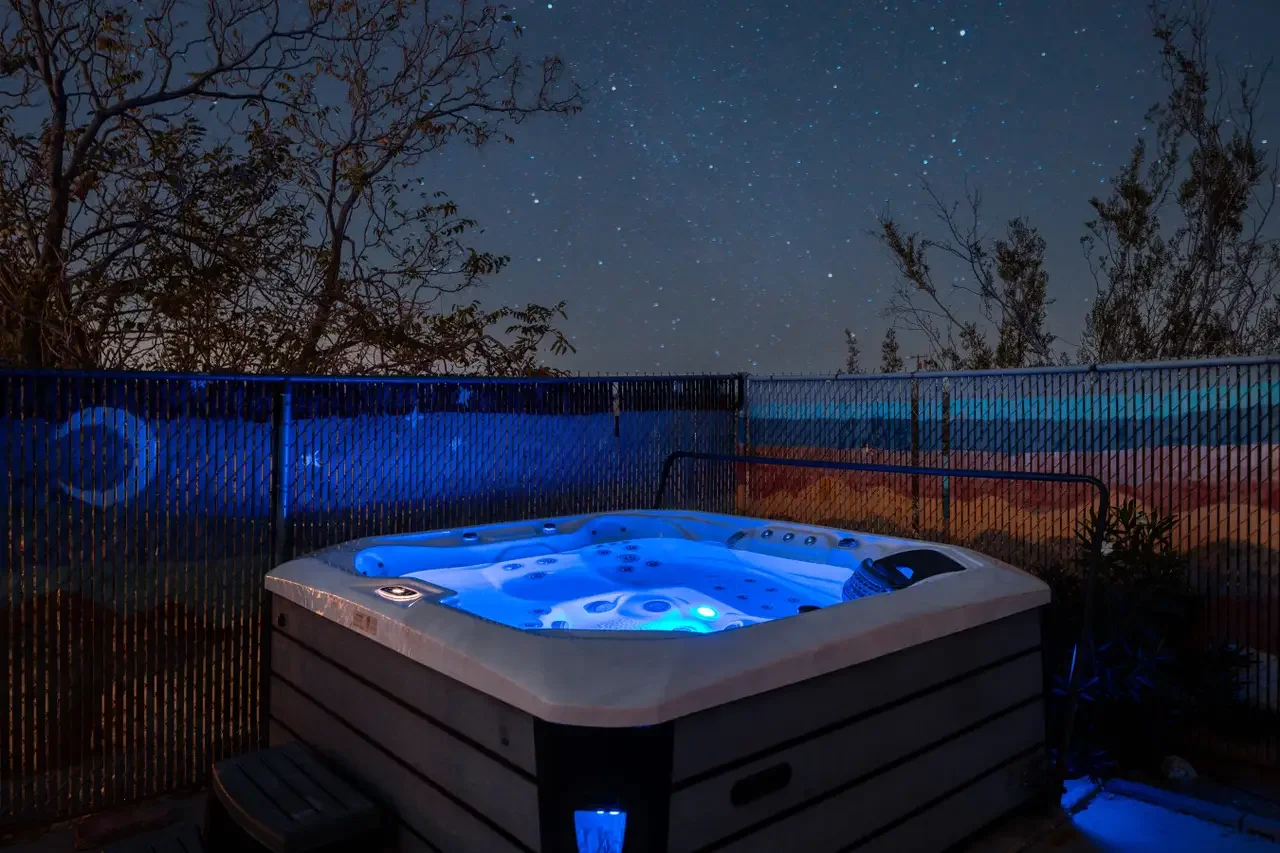 A hot tub with blue lights on a deck at night, surrounded by a metal fence and trees, under a starry sky.