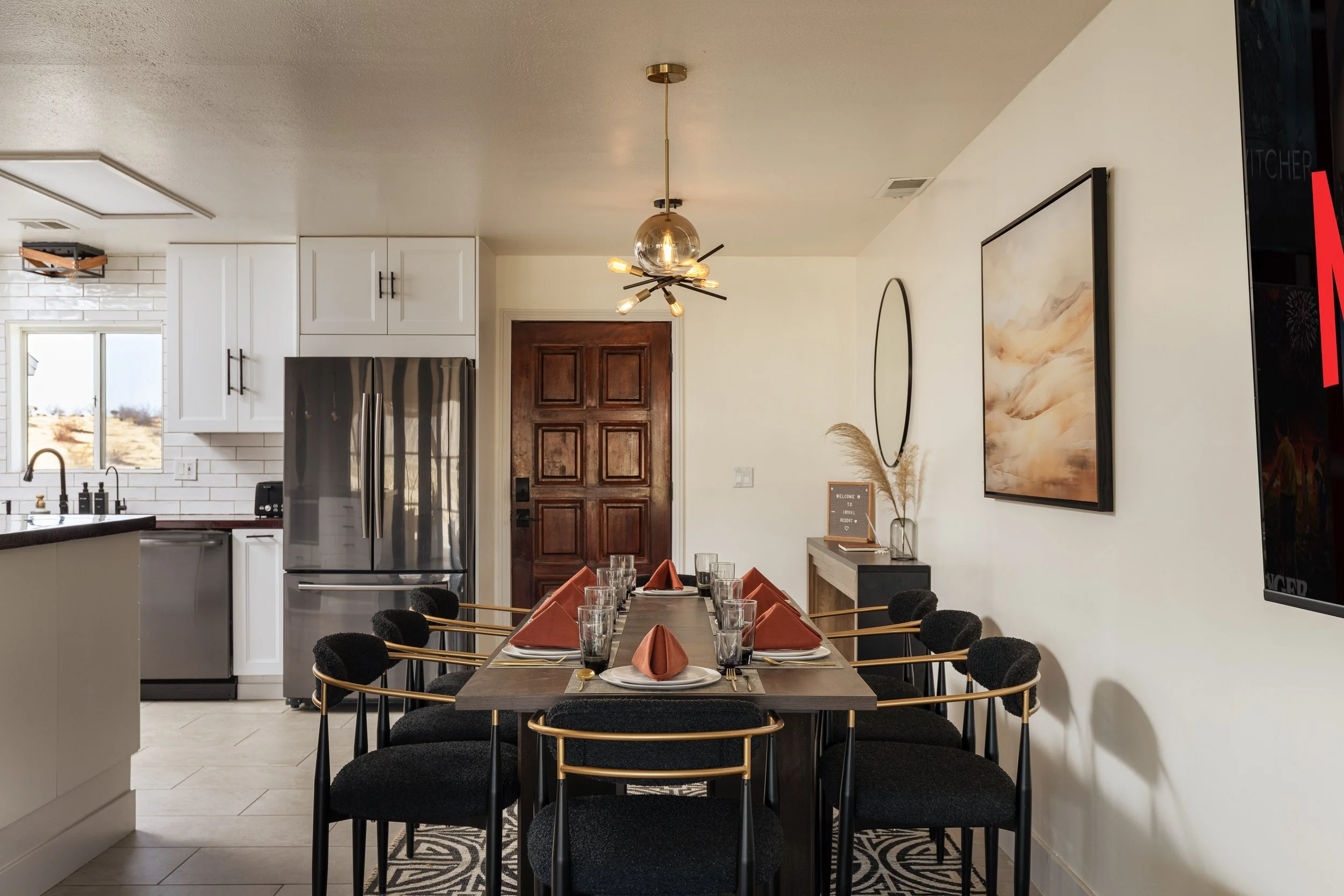Interior of a modern dining area with a long wooden table set with glasses and folded napkins, black upholstered chairs with gold frames, and a large wall-mounted artwork. The room has a white ceiling with a gold and glass chandelier, a white kitchen