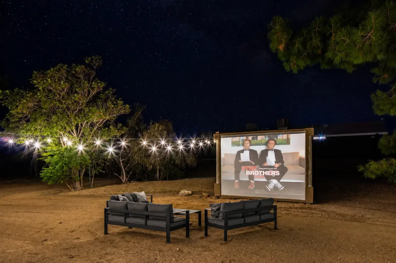 Outdoor backyard movie setup at night with a large screen showing two men in tuxedos, surrounded by trees and string lights, with chairs and a small table in front.