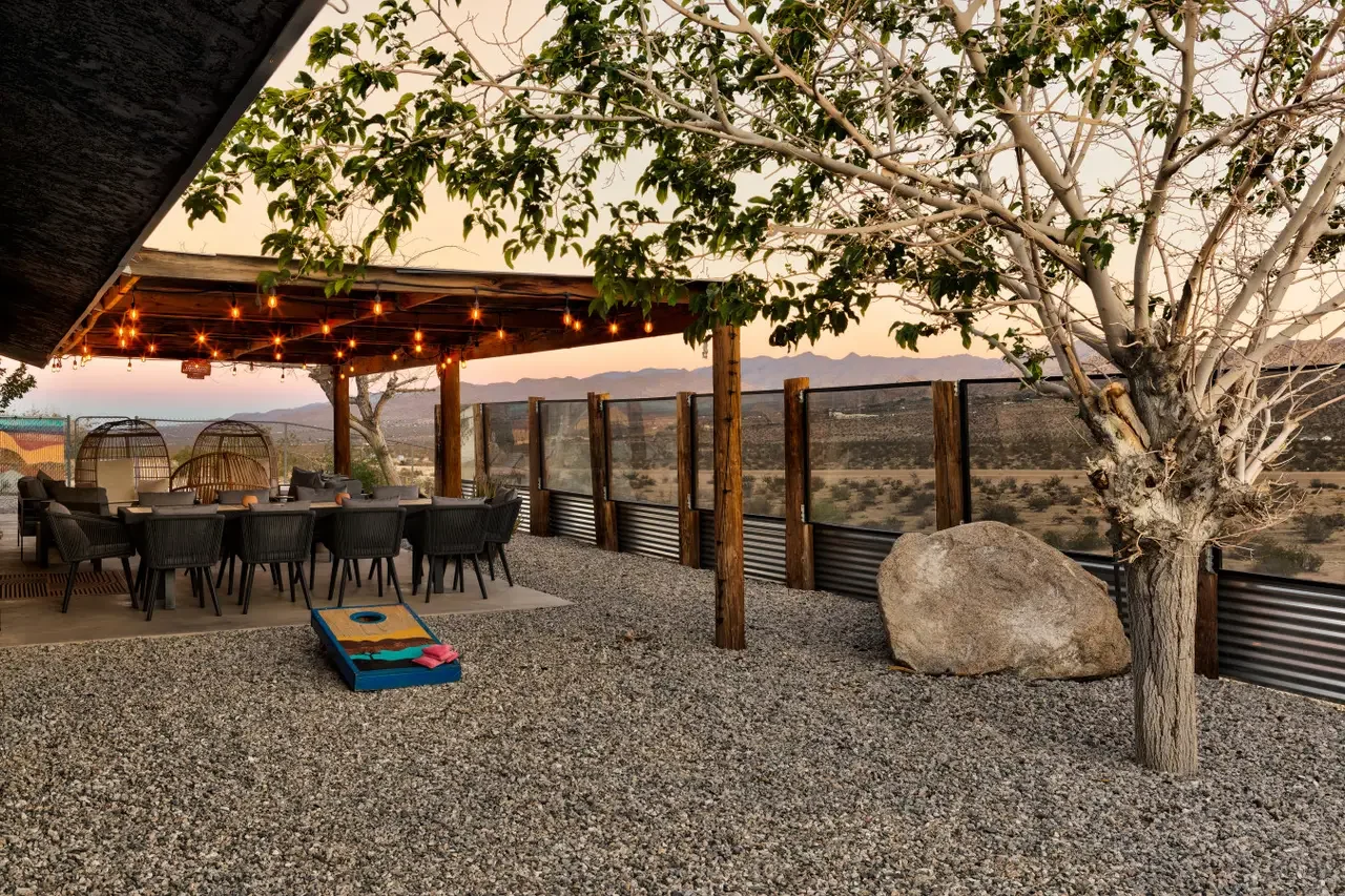 Outdoor patio area with a long dining table, black chairs, and string lights, overlooking a desert landscape with mountains in the distance, a large rock, and a leafless tree.