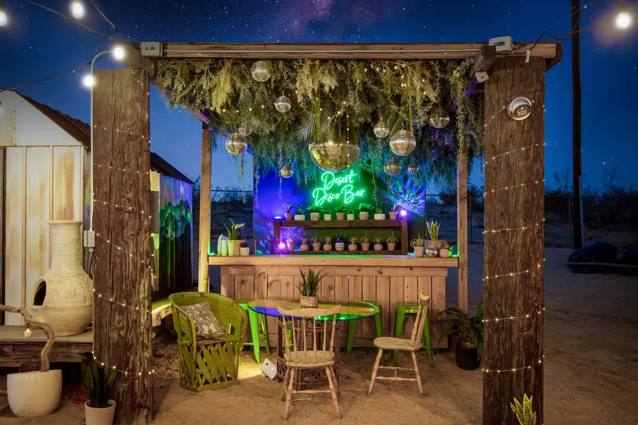 Nighttime scene of an outdoor desert disco bar with string lights, hanging disco balls, potted plants, and a neon sign that reads 'Desert Disco Bar' with a bar counter and a small table with chairs in front.