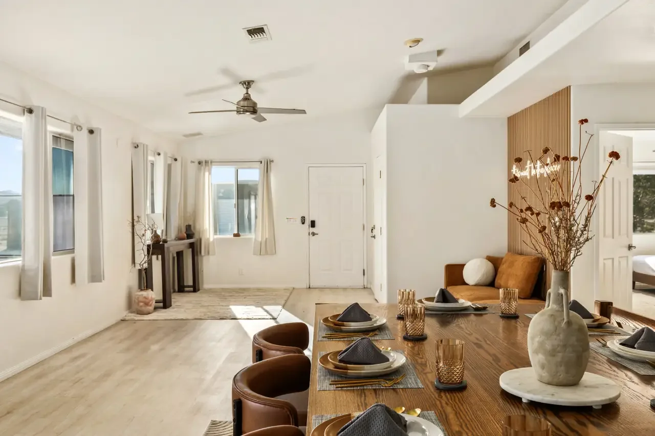 Modern dining and living room with wooden table, set for dinner, and a sitting area with cream-colored cushions, white walls, large windows with beige curtains, and a ceiling fan.